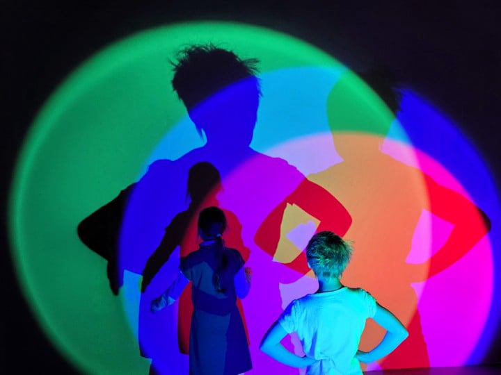 Zwei Kinder stehen im Dunklen vor einer Wand. Von hinten werden sie mit unterschiedlich farbigen Scheinwerfern beleuchtet, was bunte Schatten an die Wand wirft
