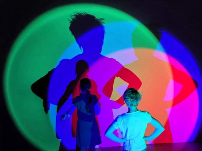 Zwei Kinder stehen im Dunklen vor einer Wand. Von hinten werden sie mit unterschiedlich farbigen Scheinwerfern beleuchtet, was bunte Schatten an die Wand wirft