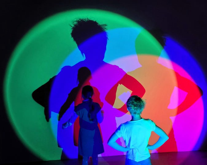 Zwei Kinder stehen im Dunklen vor einer Wand. Von hinten werden sie mit unterschiedlich farbigen Scheinwerfern beleuchtet, was bunte Schatten an die Wand wirft
